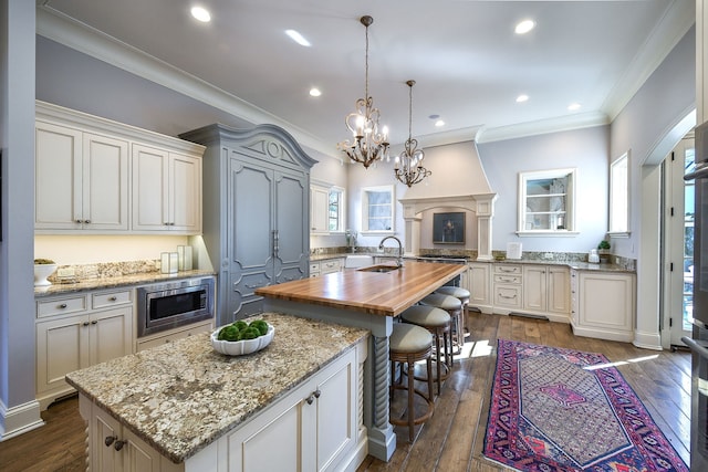 kitchen featuring an island with sink, a sink, dark wood-type flooring, wood counters, and stainless steel microwave