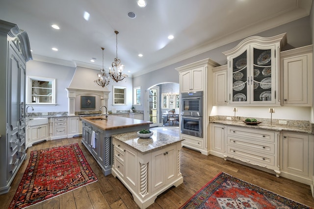 kitchen with a kitchen island with sink, glass insert cabinets, double oven, arched walkways, and dark wood-style flooring