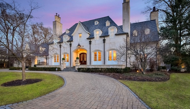 french provincial home with a high end roof, stucco siding, a chimney, curved driveway, and a lawn