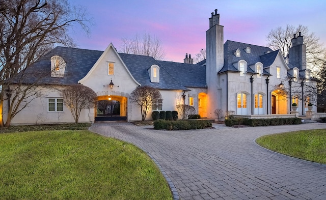 french country inspired facade featuring curved driveway, a high end roof, a lawn, stucco siding, and a chimney