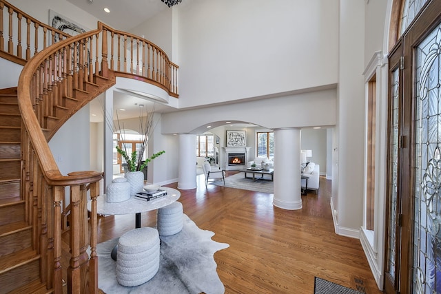 entryway featuring stairs, a lit fireplace, decorative columns, a high ceiling, and wood finished floors