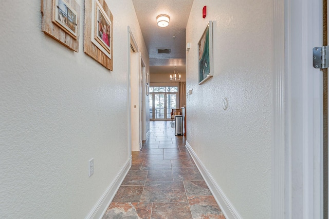 corridor featuring visible vents, stone tile flooring, an inviting chandelier, baseboards, and a textured wall