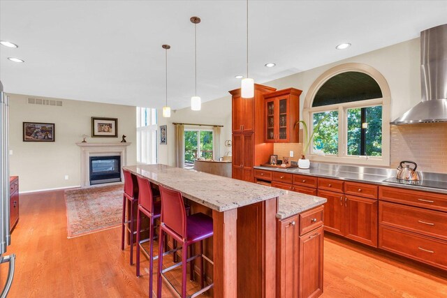 kitchen featuring stainless steel countertops, a kitchen island, a glass covered fireplace, wall chimney range hood, and black electric cooktop