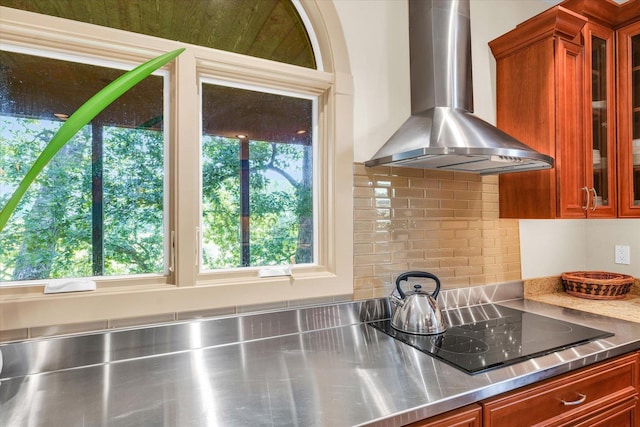 kitchen featuring backsplash, glass insert cabinets, wall chimney range hood, stainless steel countertops, and black electric cooktop