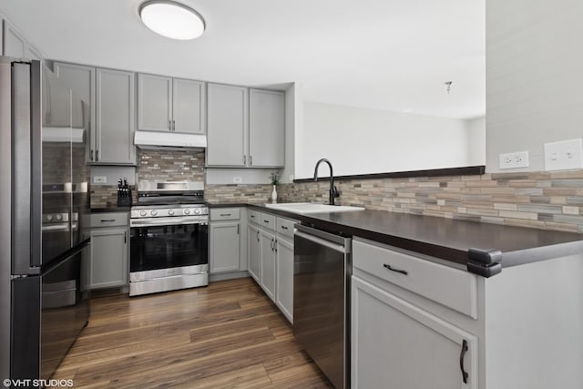 kitchen featuring a sink, under cabinet range hood, dark countertops, backsplash, and stainless steel appliances