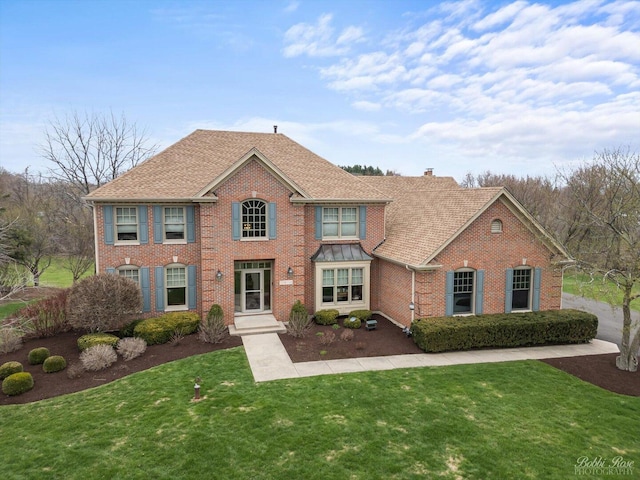 colonial-style house with roof with shingles, brick siding, a chimney, and a front lawn