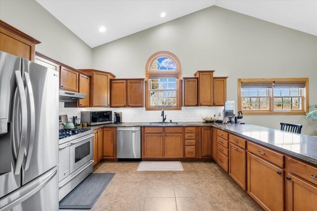 kitchen featuring high vaulted ceiling, under cabinet range hood, a sink, appliances with stainless steel finishes, and brown cabinetry