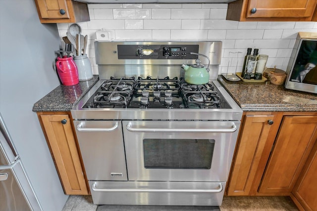 kitchen with stainless steel gas range oven, backsplash, dark stone counters, and brown cabinetry