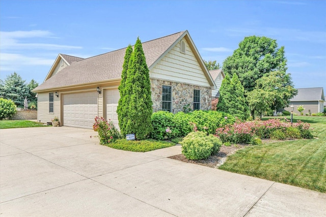 view of side of property with a yard, an attached garage, a shingled roof, concrete driveway, and stone siding