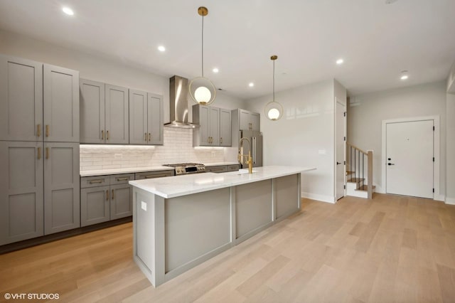 kitchen with wall chimney range hood, gray cabinets, appliances with stainless steel finishes, light wood-style floors, and a kitchen island with sink