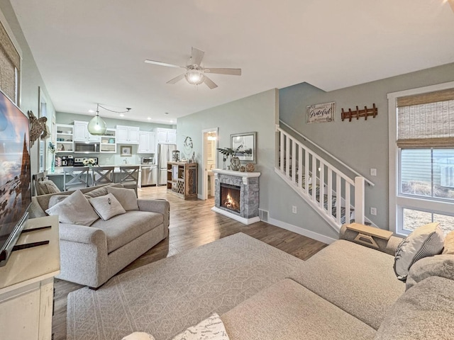 living area featuring a warm lit fireplace, wood finished floors, stairway, baseboards, and ceiling fan