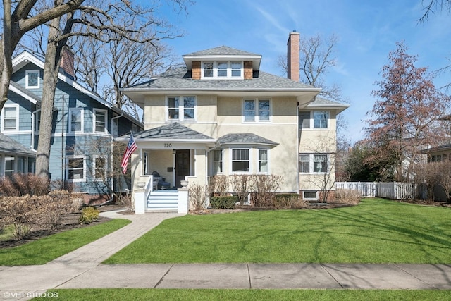 american foursquare style home with stucco siding, fence, a front yard, a shingled roof, and a chimney