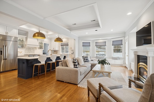 living area featuring a fireplace with flush hearth, a raised ceiling, light wood-style floors, and ornamental molding