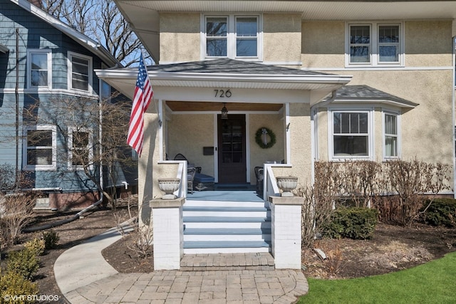 view of exterior entry featuring stucco siding, covered porch, and a shingled roof