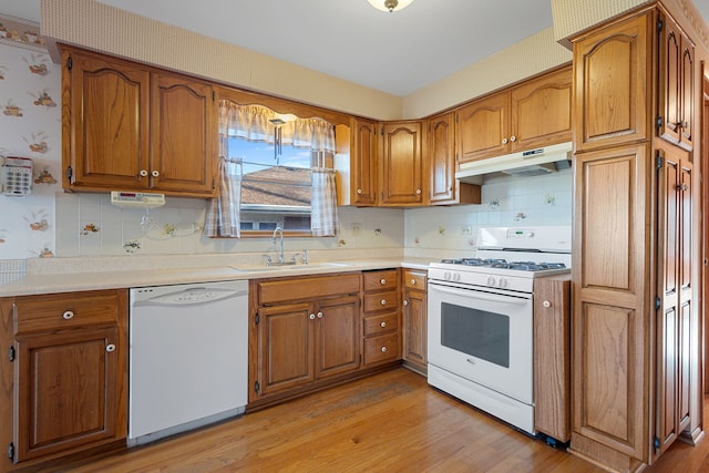 kitchen featuring wallpapered walls, under cabinet range hood, brown cabinets, white appliances, and a sink