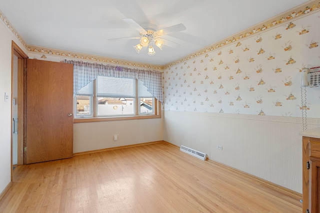 empty room featuring visible vents, wallpapered walls, light wood-type flooring, wainscoting, and a ceiling fan