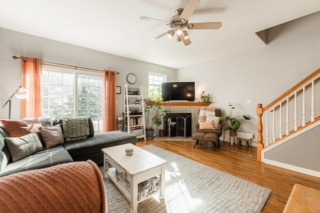 living area featuring a ceiling fan, a tiled fireplace, wood finished floors, baseboards, and stairs