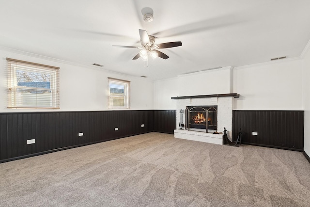 unfurnished living room featuring carpet, a ceiling fan, visible vents, wainscoting, and a brick fireplace