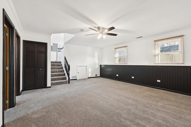 carpeted spare room featuring visible vents, crown molding, ceiling fan, stairway, and a wainscoted wall