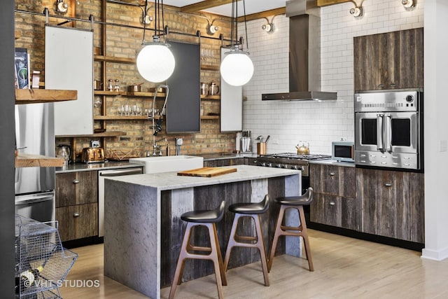 kitchen featuring backsplash, a barn door, appliances with stainless steel finishes, light wood-style floors, and wall chimney exhaust hood