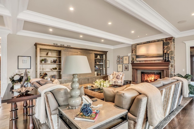 living area featuring beam ceiling, wood finished floors, a stone fireplace, crown molding, and decorative columns