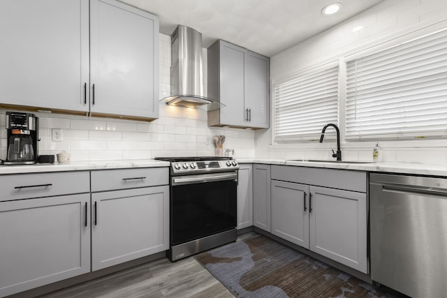 kitchen with gray cabinets, a sink, backsplash, appliances with stainless steel finishes, and wall chimney range hood