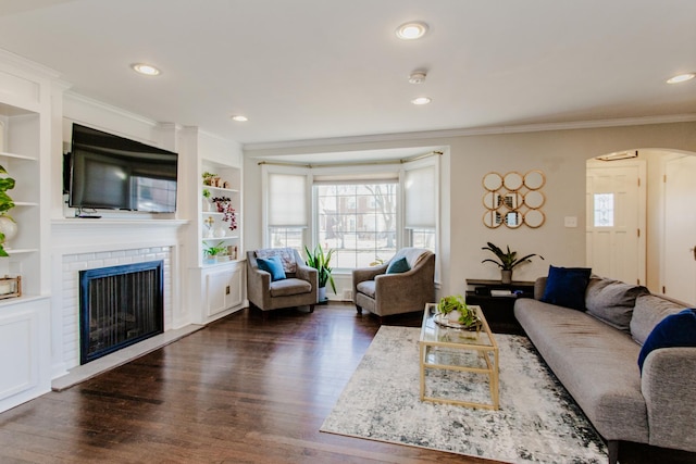 living area featuring a brick fireplace, dark wood-type flooring, crown molding, recessed lighting, and arched walkways