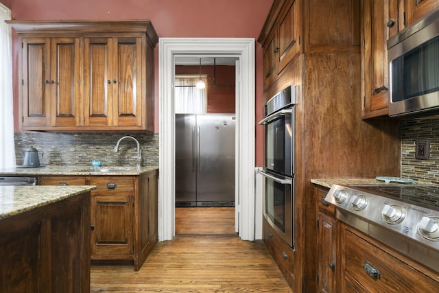 kitchen featuring light stone counters, light wood-type flooring, stainless steel appliances, decorative backsplash, and brown cabinets