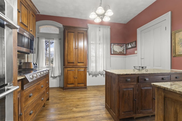 kitchen with decorative light fixtures, a wainscoted wall, light stone counters, wood finished floors, and stainless steel appliances