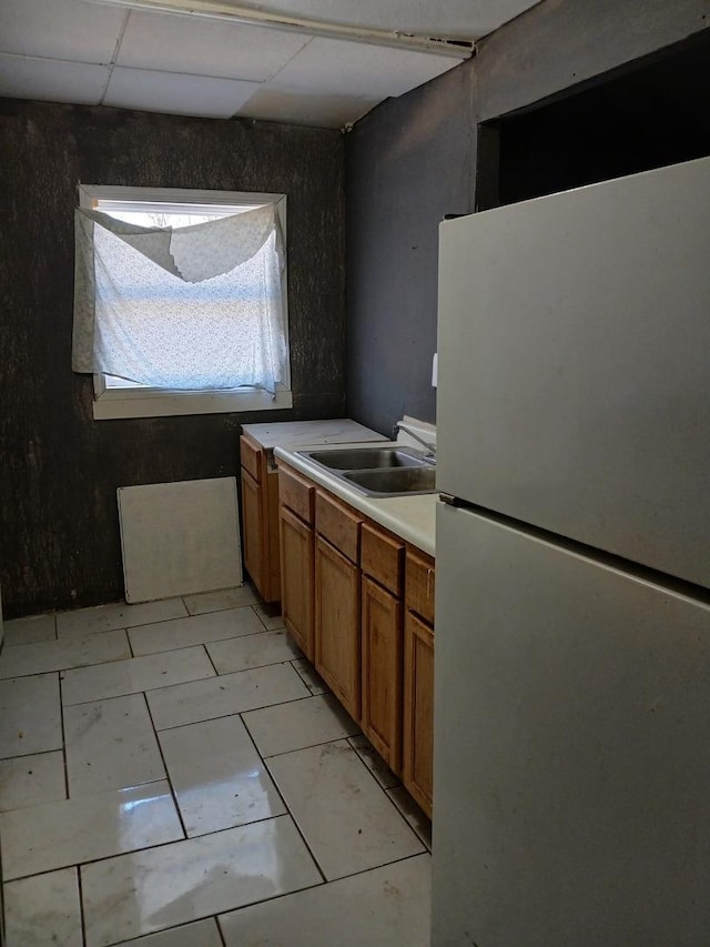 kitchen with light countertops, brown cabinets, freestanding refrigerator, a paneled ceiling, and a sink