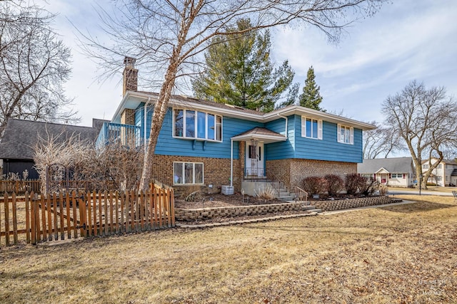 bi-level home with a front lawn, fence, cooling unit, brick siding, and a chimney