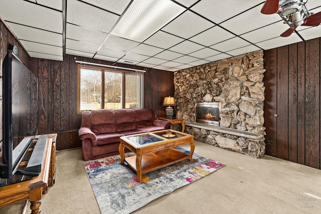 carpeted living room with a drop ceiling, wood walls, a fireplace, and a ceiling fan