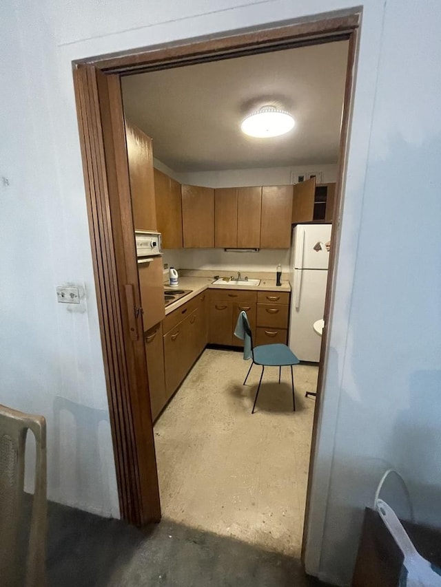 kitchen with brown cabinetry, freestanding refrigerator, a sink, light countertops, and concrete flooring