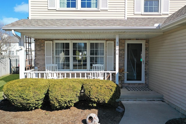 view of exterior entry with a porch, fence, brick siding, and a shingled roof