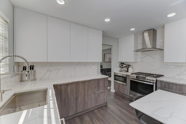 kitchen featuring a sink, wall chimney range hood, light stone counters, stainless steel appliances, and modern cabinets