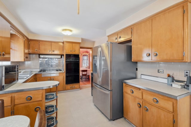 kitchen with under cabinet range hood, appliances with stainless steel finishes, brown cabinetry, light countertops, and decorative backsplash