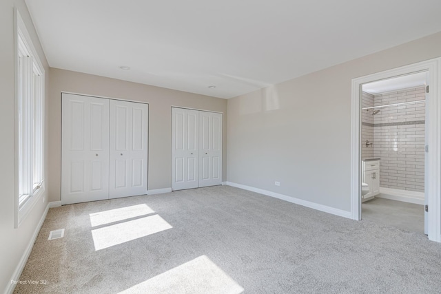 unfurnished bedroom featuring baseboards, visible vents, carpet floors, ensuite bathroom, and two closets