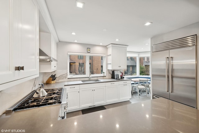 kitchen with white cabinets, built in refrigerator, wall chimney range hood, and a sink