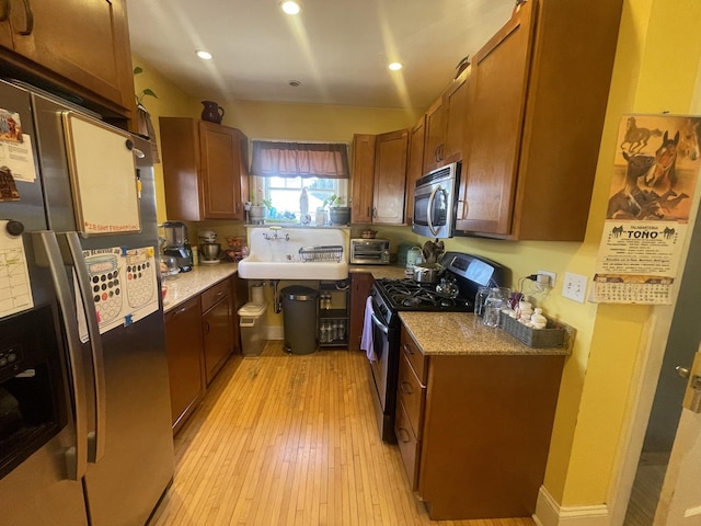 kitchen with light wood-style flooring, a sink, appliances with stainless steel finishes, brown cabinetry, and light stone countertops