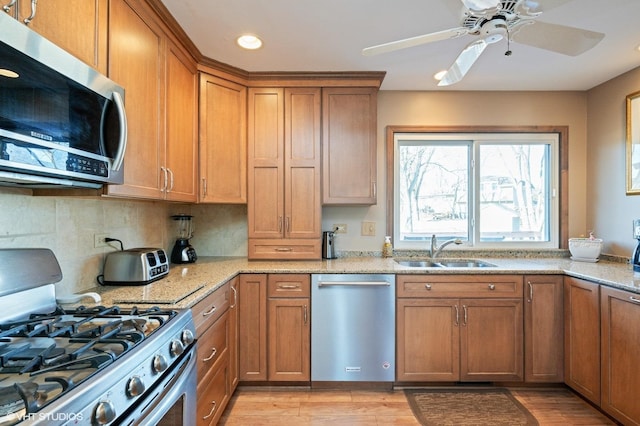 kitchen with light wood-type flooring, a sink, appliances with stainless steel finishes, brown cabinetry, and light stone countertops