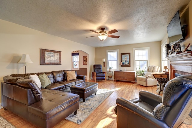 living room featuring a ceiling fan, a textured ceiling, arched walkways, light wood-style floors, and a brick fireplace