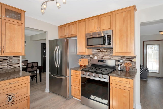 kitchen with backsplash, glass insert cabinets, light wood-style flooring, appliances with stainless steel finishes, and dark stone countertops