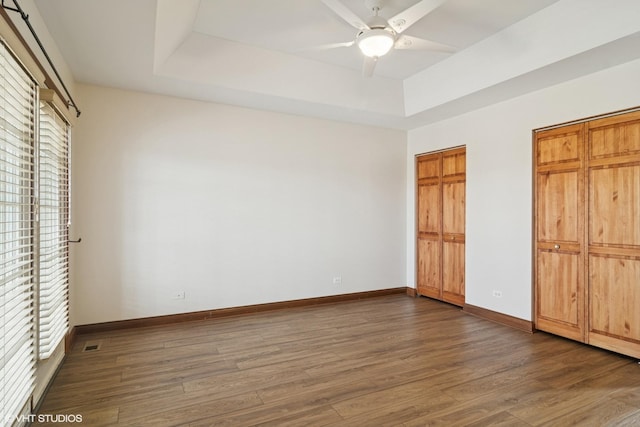 unfurnished bedroom featuring baseboards, multiple closets, a raised ceiling, a ceiling fan, and dark wood-style flooring