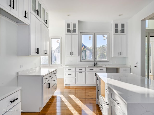 kitchen with glass insert cabinets, light stone countertops, light wood-type flooring, white cabinetry, and a sink