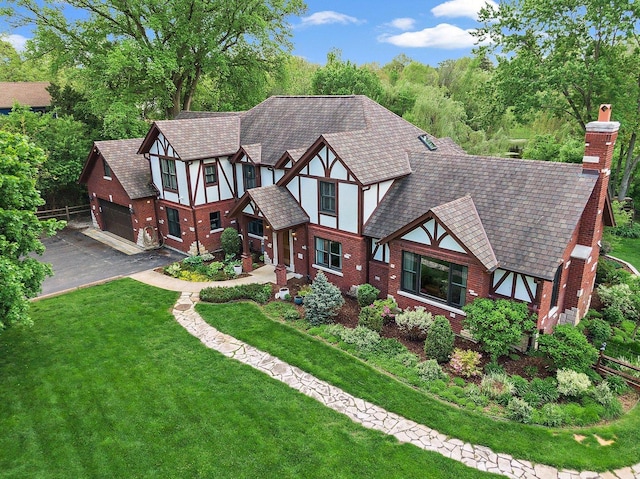 tudor-style house with brick siding, a front lawn, a chimney, a garage, and driveway