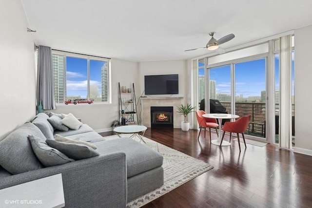 living room featuring a wall of windows, a ceiling fan, wood finished floors, baseboards, and a lit fireplace