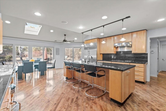 kitchen with under cabinet range hood, gas cooktop, built in fridge, light wood-style floors, and decorative backsplash