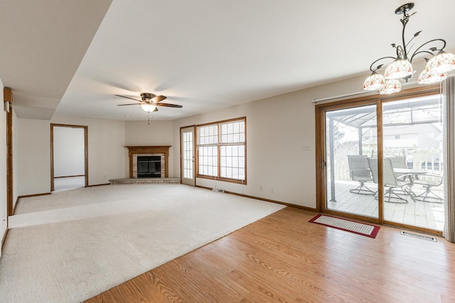 unfurnished living room featuring plenty of natural light, a brick fireplace, wood finished floors, and ceiling fan with notable chandelier