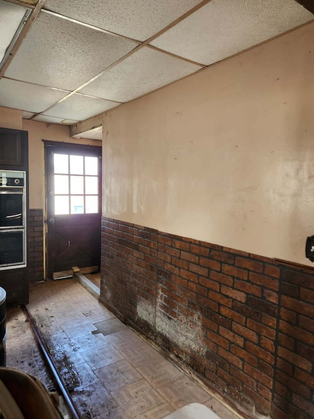 interior space featuring stainless steel double oven, a paneled ceiling, brick wall, and wainscoting