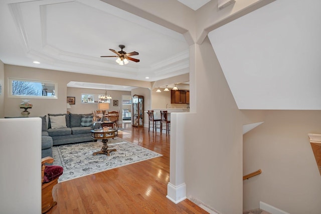 living area with baseboards, a tray ceiling, light wood-style flooring, ornamental molding, and ceiling fan with notable chandelier